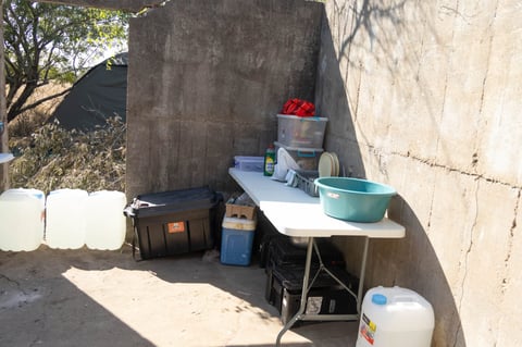 Outdoor kitchen setup against concrete walls with folding table, stove, sink, and storage containers in sandy area