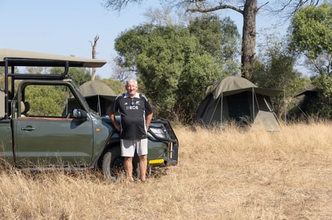 Person standing beside open-door safari vehicle in dry grassland with canvas tents and trees in background