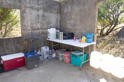 Outdoor kitchen setup under concrete structure with coolers, portable stove, water containers, and storage shelves